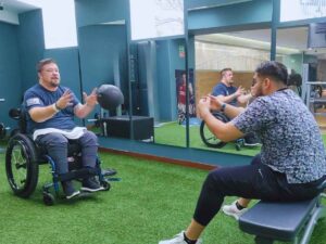 Paul, seated in his wheelchair, joyfully throws a ball to a physical therapist as part of his recovery program from epidural stimulation treatment for spinal cord injury, showcasing an interactive therapy session.