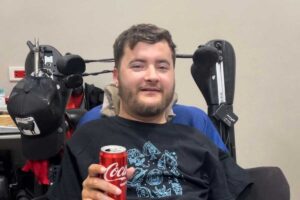 Quadriplegic patient Bryce, C5 complete smiling and holding a can of Coca-Cola, showcasing the progress made with Verita Neuro’s epidural stimulation treatment. The image highlights Bryce’s improved upper body strength and motor function as he is able to squeeze the can.