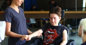 Alissa, a C4 quadriplegic patient, undergoing physical therapy with the assistance of a therapist, focusing on arm movement exercises. She is seated in a wheelchair, determined and focused on her rehabilitation