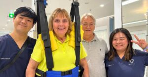 Dominique, a determined C6 quadriplegic patient, stands in a supportive harness during a therapy session, assisted by two Verita Neuro specialists and supported by her husband. The bright and modern therapy room reflects the advanced care and positivity driving her recovery through Epidural Stimulation and rehabilitation.