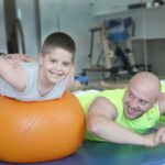 A young boy with Cerebral Palsy smiling during physical therapy on an orange exercise ball, supported by a therapist and accompanied by a parent, highlighting rehabilitation and progress.