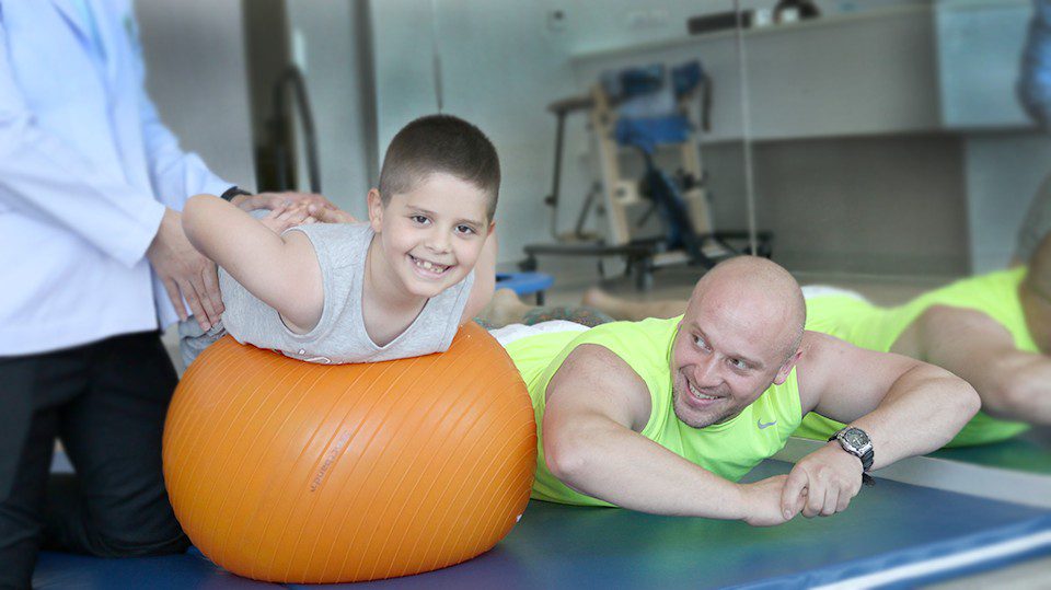 A young boy with Cerebral Palsy smiling during physical therapy on an orange exercise ball, supported by a therapist and accompanied by a parent, highlighting rehabilitation and progress.