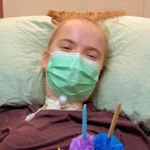 Makayla, a 16-year-old C3 complete quadriplegic patient, smiles from her hospital bed at Verita Neuro in Thailand, where she is receiving Epidural Stimulation treatment. She wears a green surgical mask and maroon top, with a visible tracheostomy at her neck. She holds colorful paper flowers and a candle from a celebration held during her recovery journey.