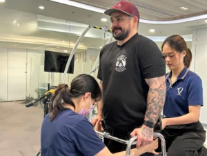 Tylan, a male T6 Complete Spinal Cord Injury Patient, with a full sleeve tattoo, performing excercises with female Physical Therapist at Verita Neuro in Thailand, following Treatment with Stem Cells, Epidural Stimulation and LamiSpine