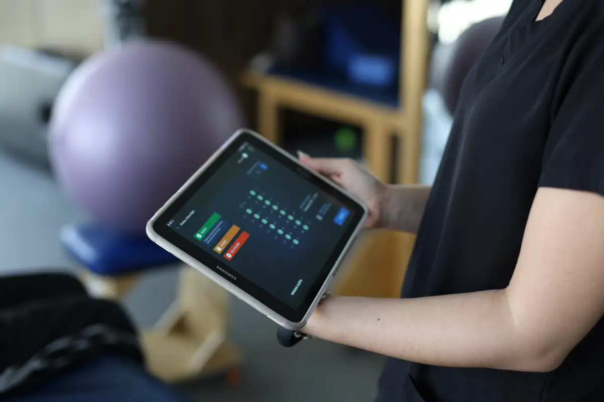 Mapping Specialist is seen programming an Epidural Stimulator on a Tablet, during a Physical Therapy session. The therapist holds the tablet in a room filled with physical therapy equipment including a purple yoga ball.