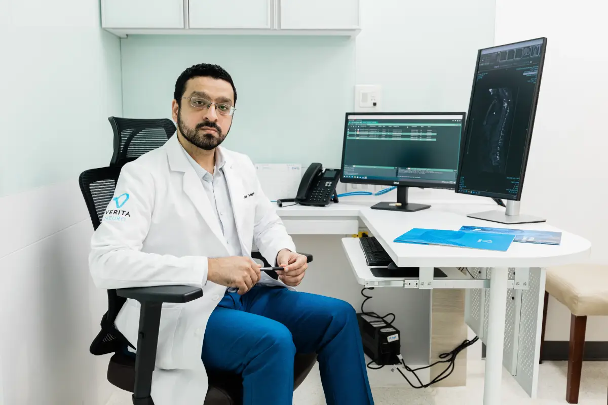 Global Medical Director Dr. Nasir Majeed in scrubs sitting at a desk with computers displaying medical data, facing the camera